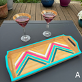 A rectangular bamboo serving tray with a turquoise painted border and a colorful chevron pattern in pink, teal, yellow, and white on the top. Two stemmed glasses with pink drinks sit behind it on a dark table, with an outdoor deck and potted plants in the background.