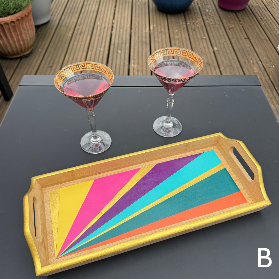 A rectangular bamboo serving tray with colorful painted geometric stripes in yellow, pink, purple, teal, and orange, styled on a dark table next to two martini glasses on a wooden deck.