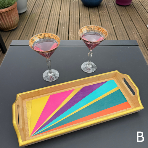 A rectangular bamboo serving tray with colorful painted geometric stripes in yellow, pink, purple, teal, and orange, styled on a dark table next to two martini glasses on a wooden deck.