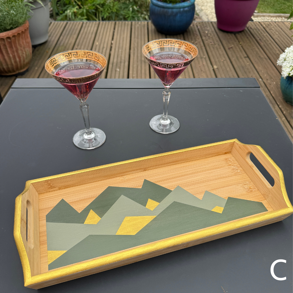 A rectangular bamboo serving tray with integrated handles, featuring a painted geometric mountain pattern in olive green, gray, and yellow. The tray is shown on a dark gray table outdoors, with two martini glasses in the background.