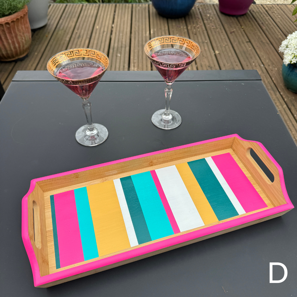 A rectangular wooden serving tray with bright pink handles and a striped interior in pink, teal, yellow, white, and teal, shown on a dark table outdoors with two martini glasses behind it.