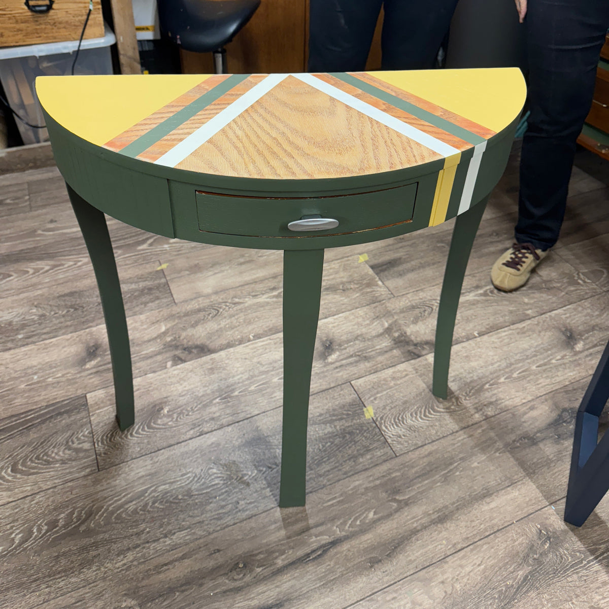 A green half-moon console table with a decorative geometric wood inlay on the top, shown in a workshop with two people standing behind it.