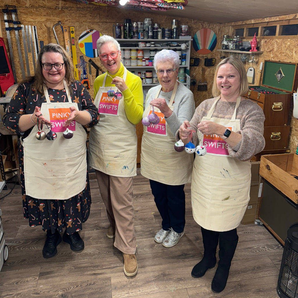 Four women wearing beige aprons stand together in a decorated craft room, each holding painted ceramic christmas baubles