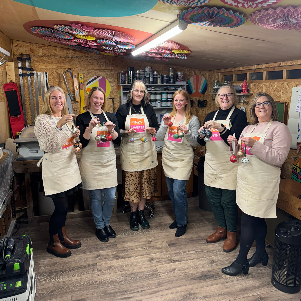 Six women stand together in a colorful craft room, holding up handpainted christmas cermic baubles after a pinkyswifts christmas workshop