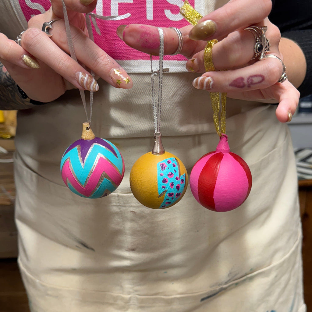 Three hand-painted Christmas ornaments in pink, yellow, and blue are held up by a person wearing a beige apron with 'PINKY SKIETS' printed on it, in what appears to be a craft or paint studio.