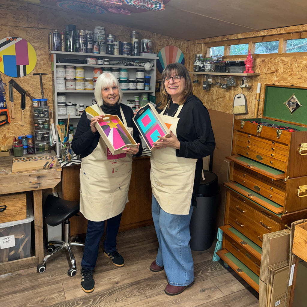 Two women in a workshop holding colorful art pieces.