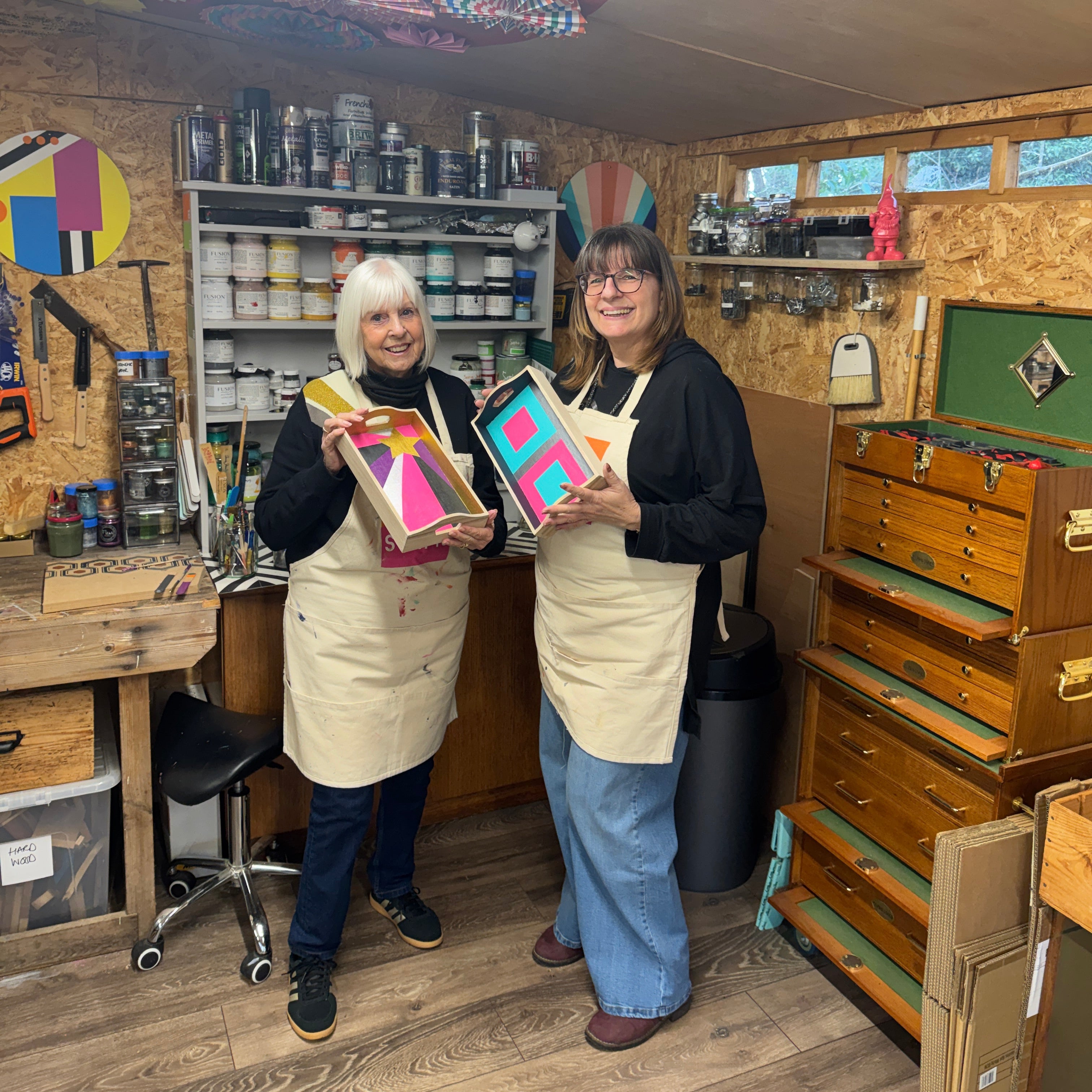 Two women in a workshop holding colorful art pieces.