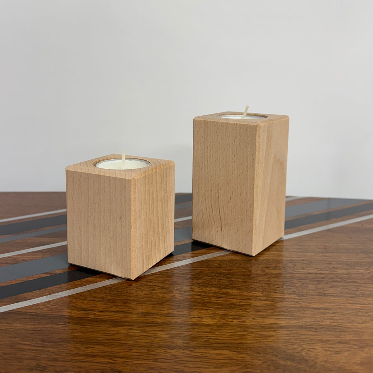 Two square wooden candle holders with tea light candles, displayed on a polished wooden table against a plain light gray wall.