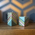 Two decorative square candles with a pastel chevron pattern in mint green, dusty pink, and metallic gold, displayed on a wooden surface against a geometric patterned wall.