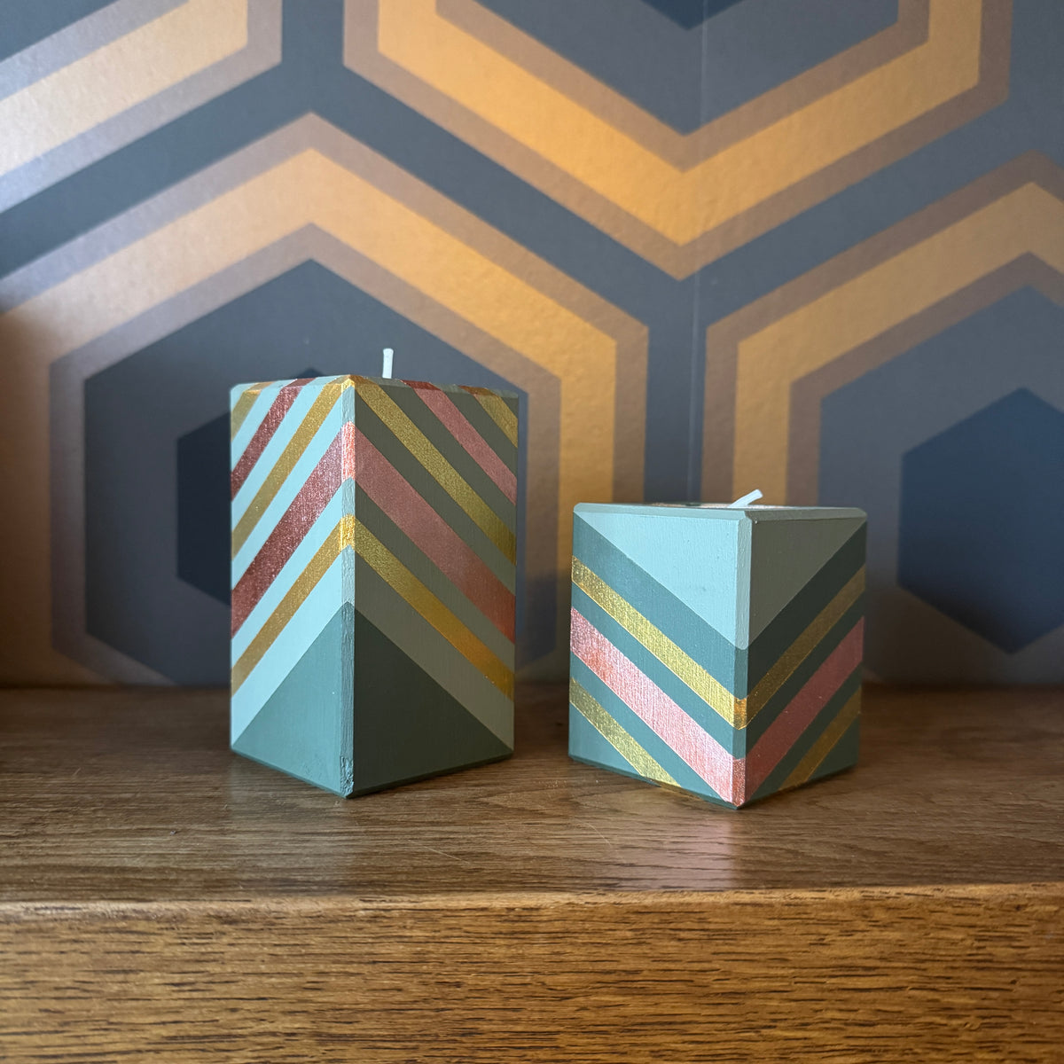 Two decorative square candles with a pastel chevron pattern in mint green, dusty pink, and metallic gold, displayed on a wooden surface against a geometric patterned wall.