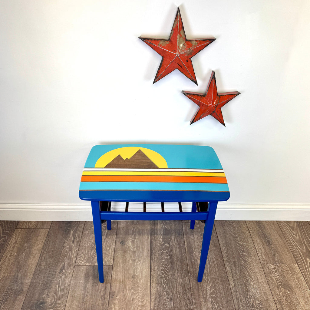 A small mid century table with a colorful retro painted top design of mountains and a sun, set on a wood floor against a white wall with two red star decorations.