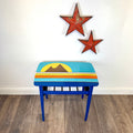 A small mid century table with a colorful retro painted top design of mountains and a sun, set on a wood floor against a white wall with two red star decorations.