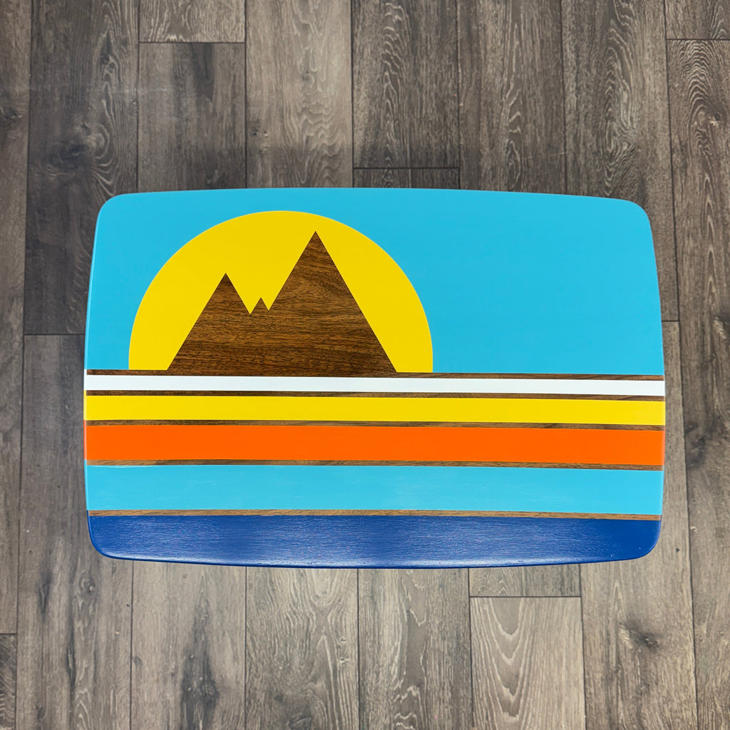 A top-down view of a wooden mid century table with a colorful retro sunset and mountain design, placed on a dark wood plank floor. 