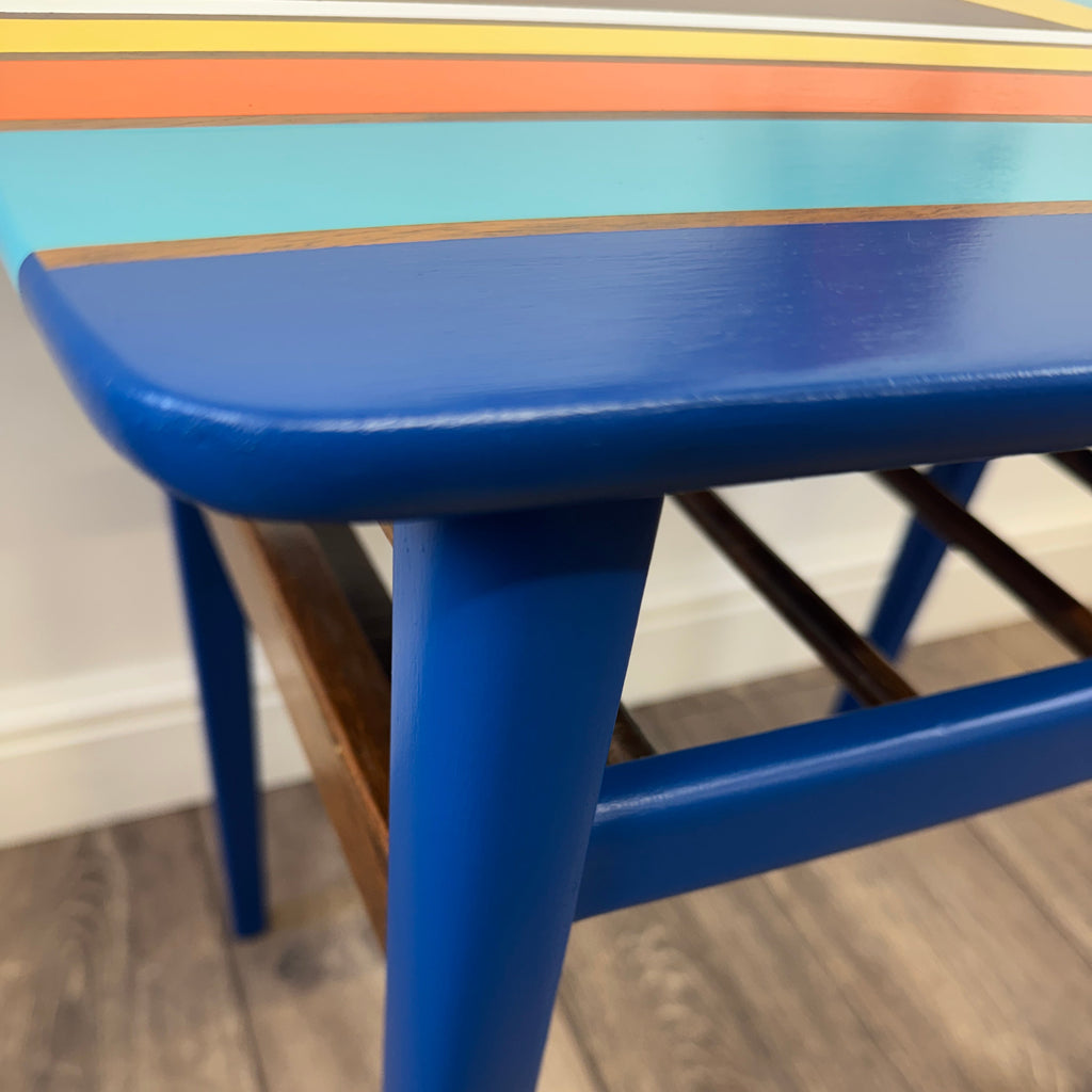 A close-up of a vibrant blue wooden mid century table with colorful retro painted stripes on the top, set on a gray wood-look floor.