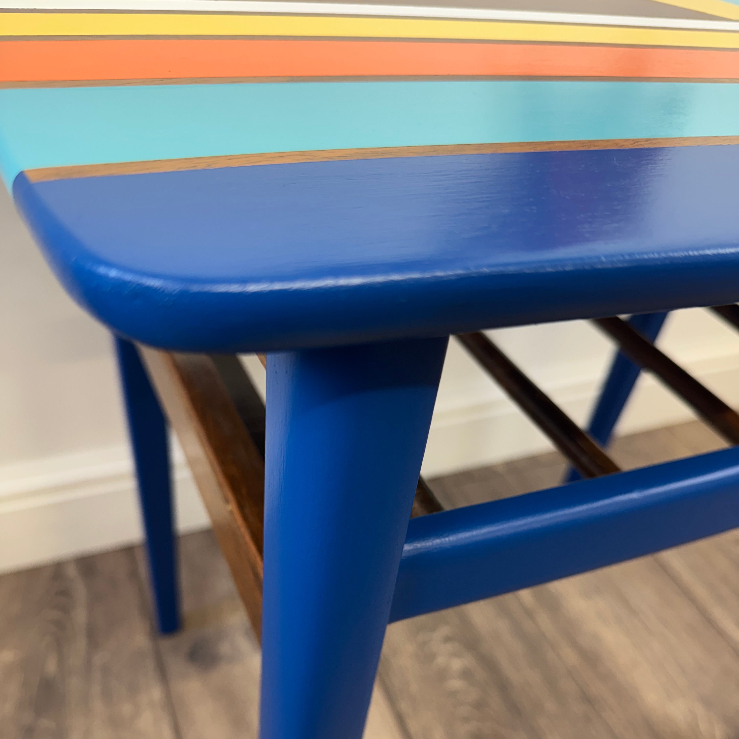 A close-up of a vibrant blue wooden mid century table with colorful retro painted stripes on the top, set on a gray wood-look floor.