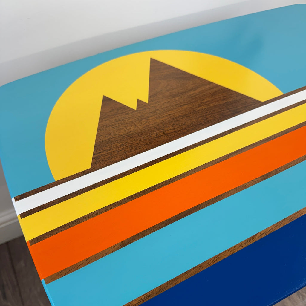 A close-up of a colorful mid century table featuring a retro sunset mountain design with wood-grain accents, set in a room with a gray wall and wood floor.