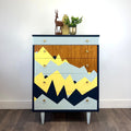 A mid-century modern chest of drawers with a geometric mountain pattern in yellow, navy, light blue, and wood grain, styled in a room with a white wall and wood floor.