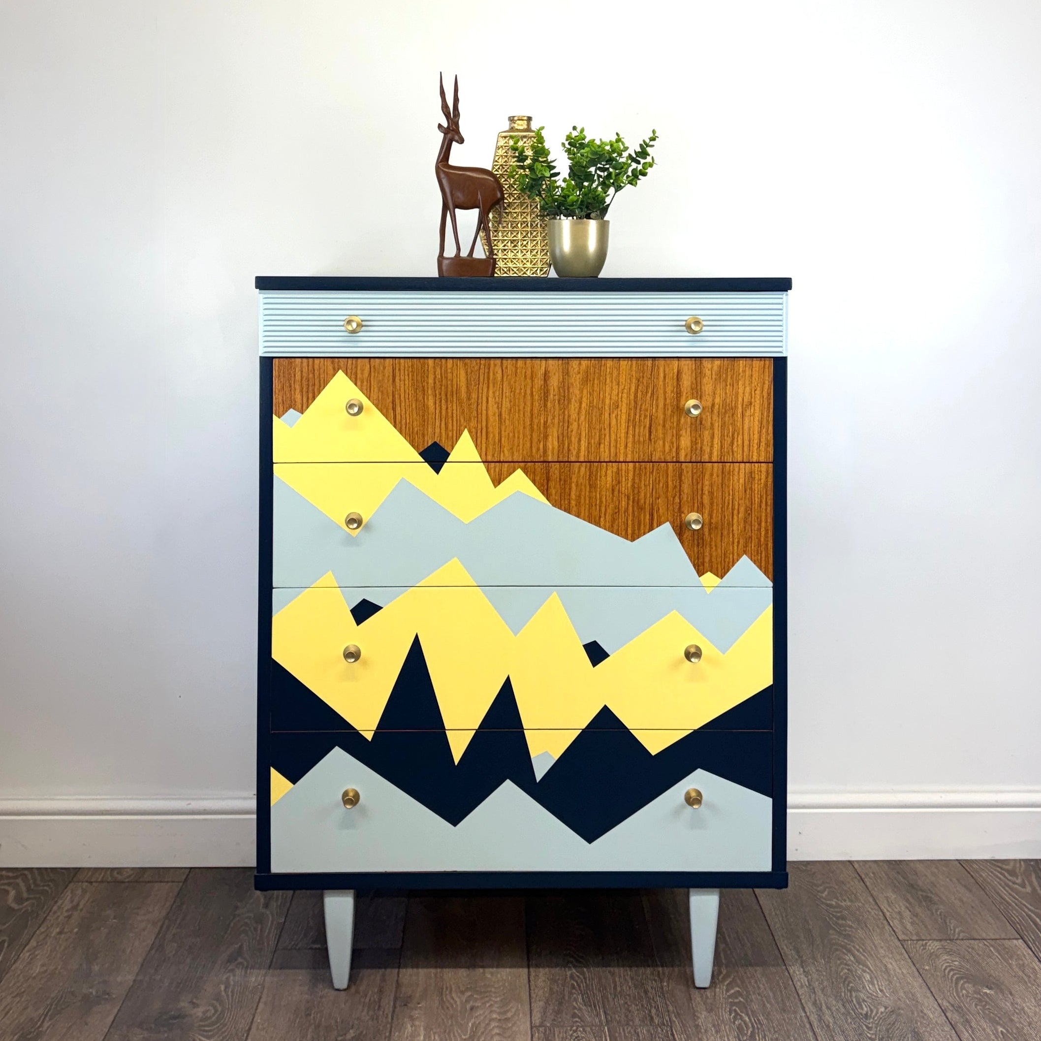 A mid-century modern chest of drawers with a geometric mountain pattern in yellow, navy, light blue, and wood grain, styled in a room with a white wall and wood floor.