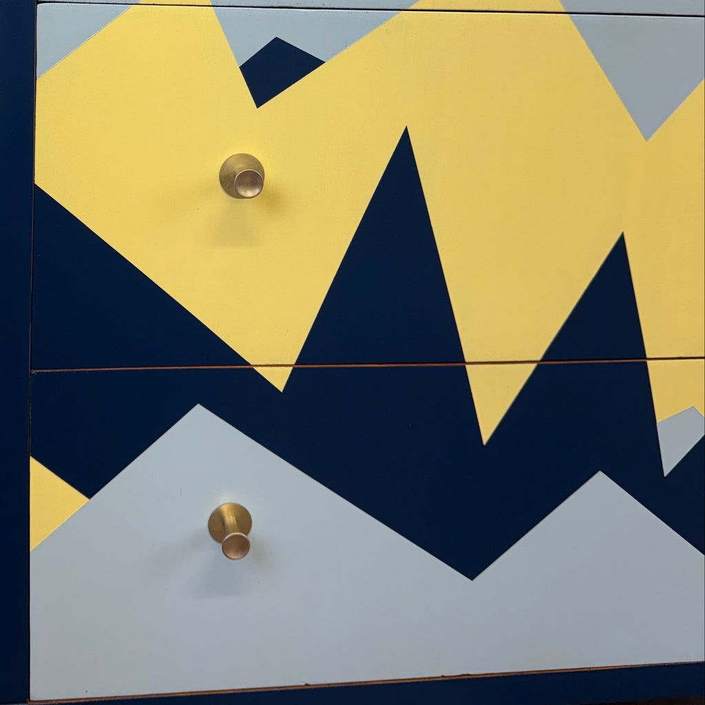 A close-up of a dresser with a geometric mountain pattern in yellow, light blue, and navy blue, featuring round brass drawer pulls, set against a white wall and wood floor.