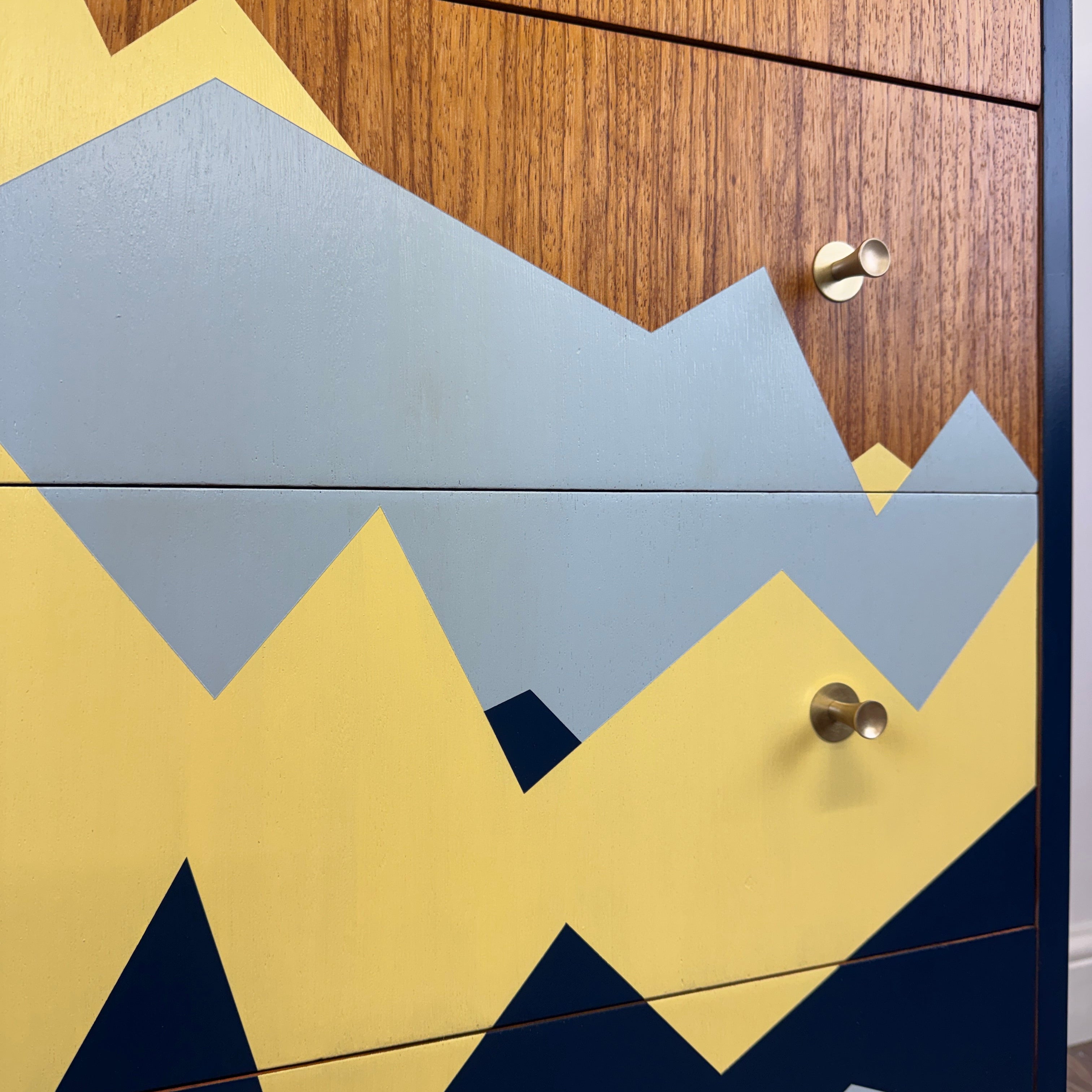 Close-up of a modern dresser or cabinet with a geometric mountain pattern in yellow, slate blue, and navy on a wood-grain front, with brass drawer pulls.