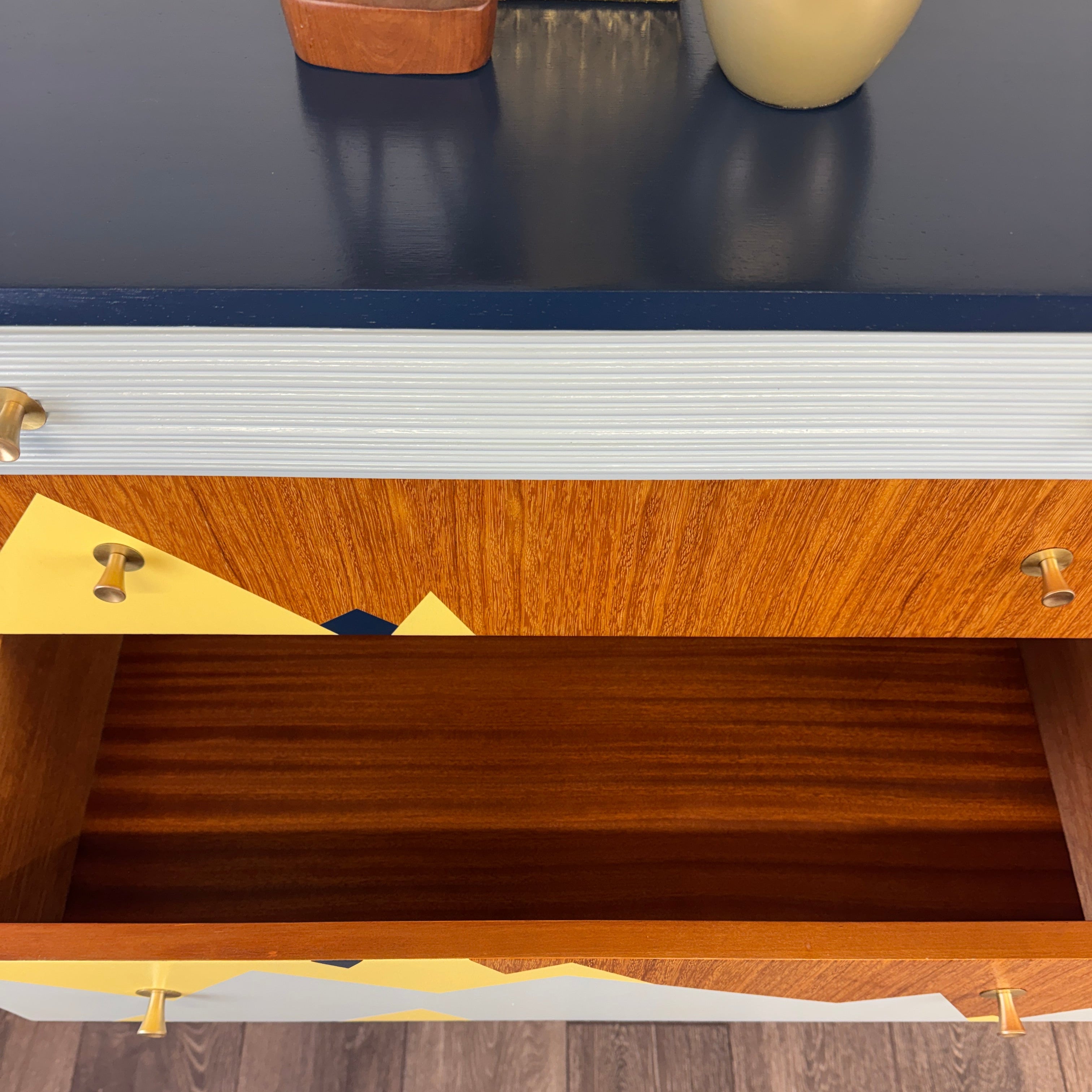 An open wooden drawer of a mid-century modern credenza or sideboard, showing a warm brown wood interior and brass handles, set against a dark blue top surface and light wood flooring.