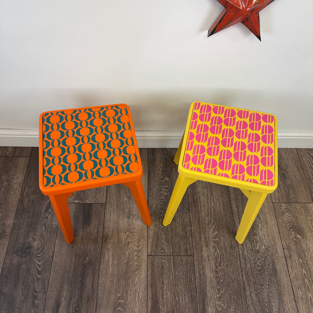 Two colorful metal stools on a wood-look floor against a white wall. The left stool is orange with a teal geometric pattern on the seat, and the right stool is yellow with a hot pink geometric pattern on the seat.