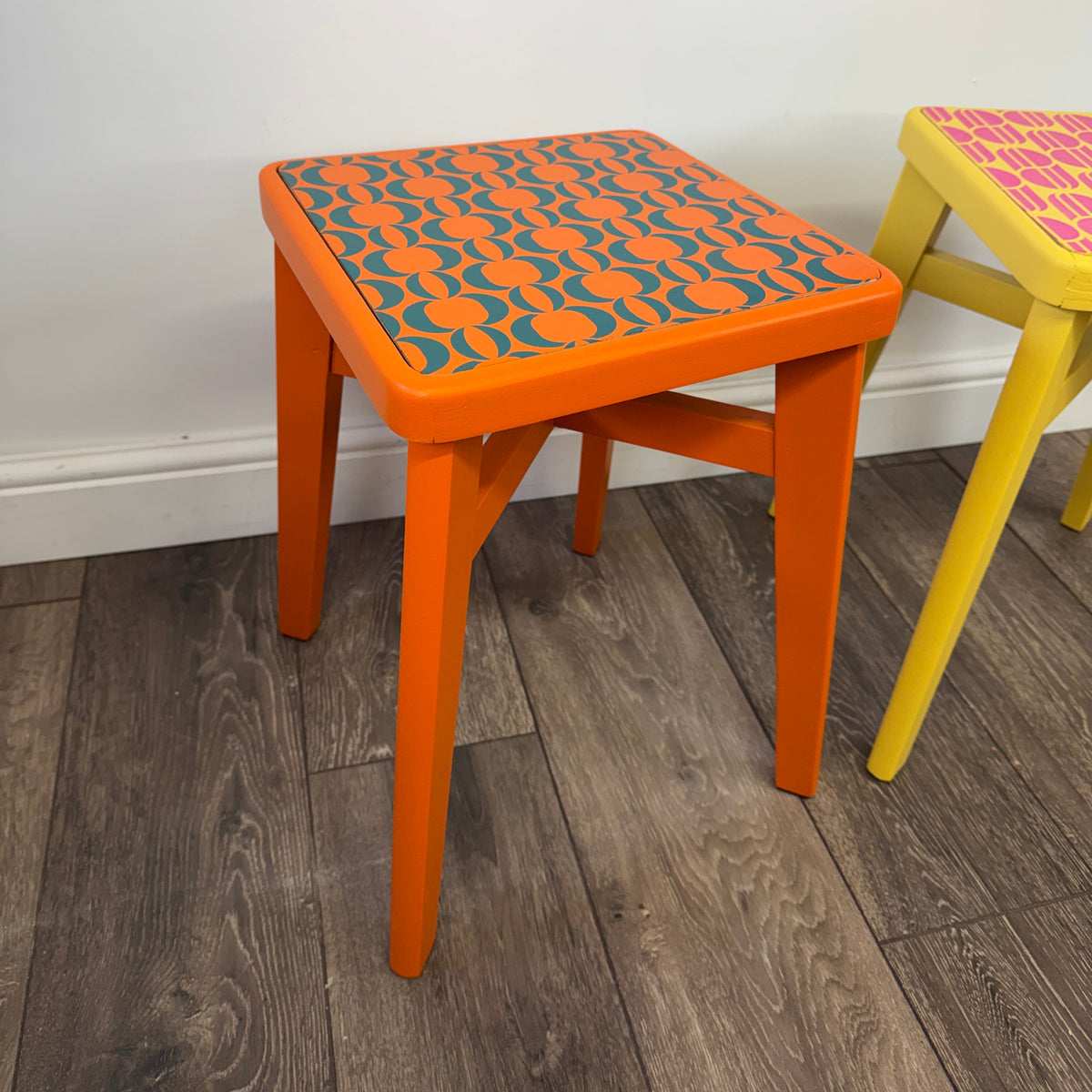 An orange wooden stool with a patterned orange and teal seat, shown next to another yellow stool on a wood floor against a white wall.