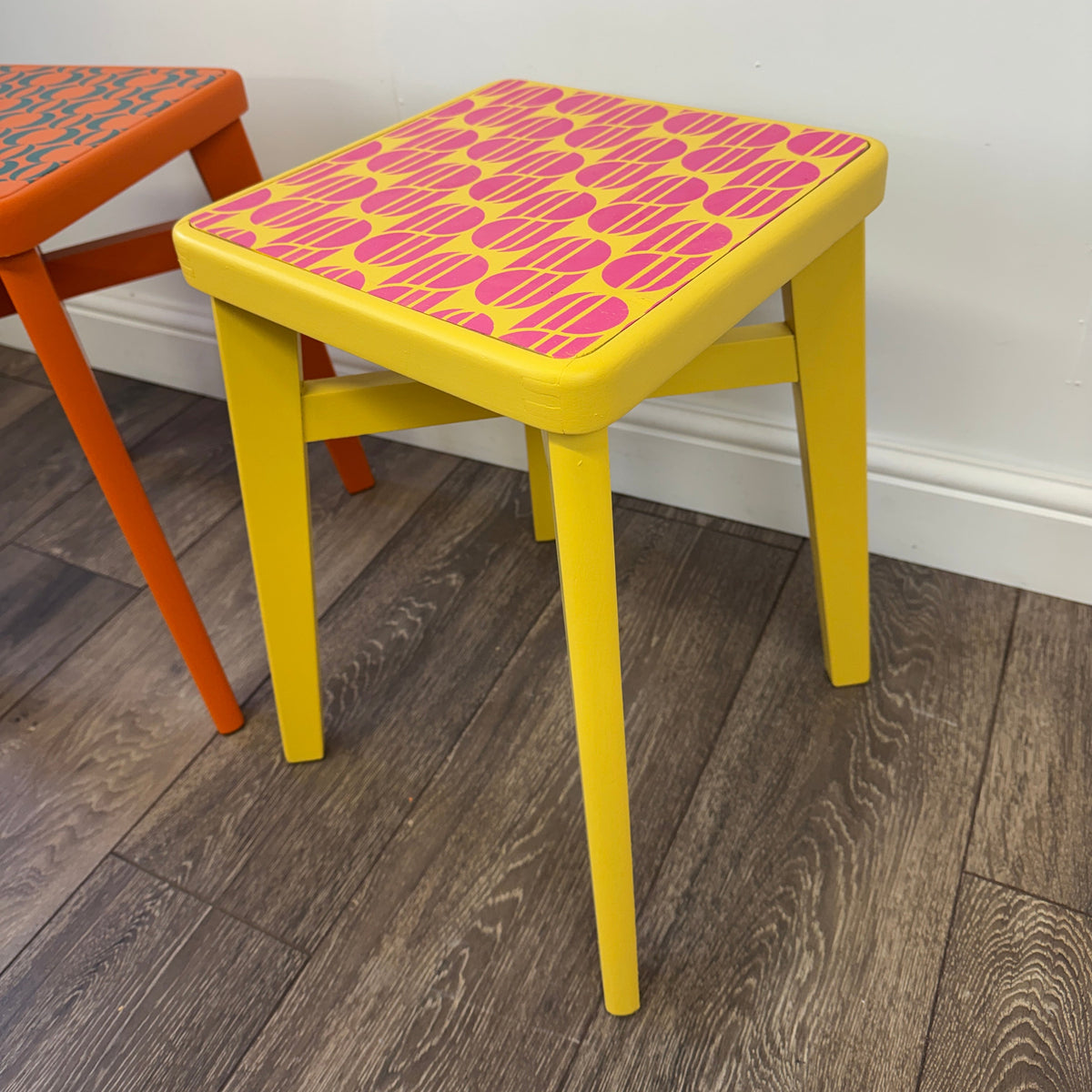 A square yellow stool with a pink and orange patterned seat, shown in a room with a white wall and wood-look floor. Another stool is partially visible on the left.