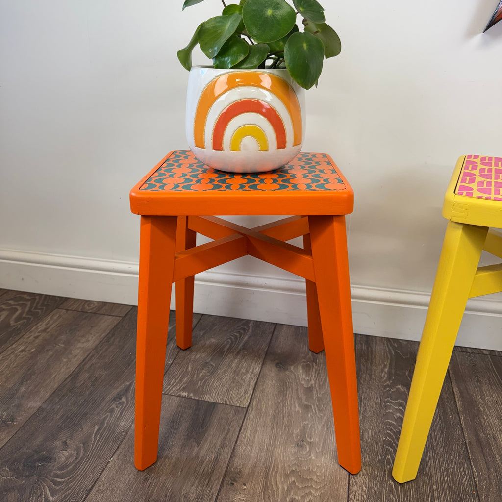An orange wooden stool with a patterned top holding a potted green plant in a white pot with an orange and yellow sunburst design, set against a white wall and dark wood floor.