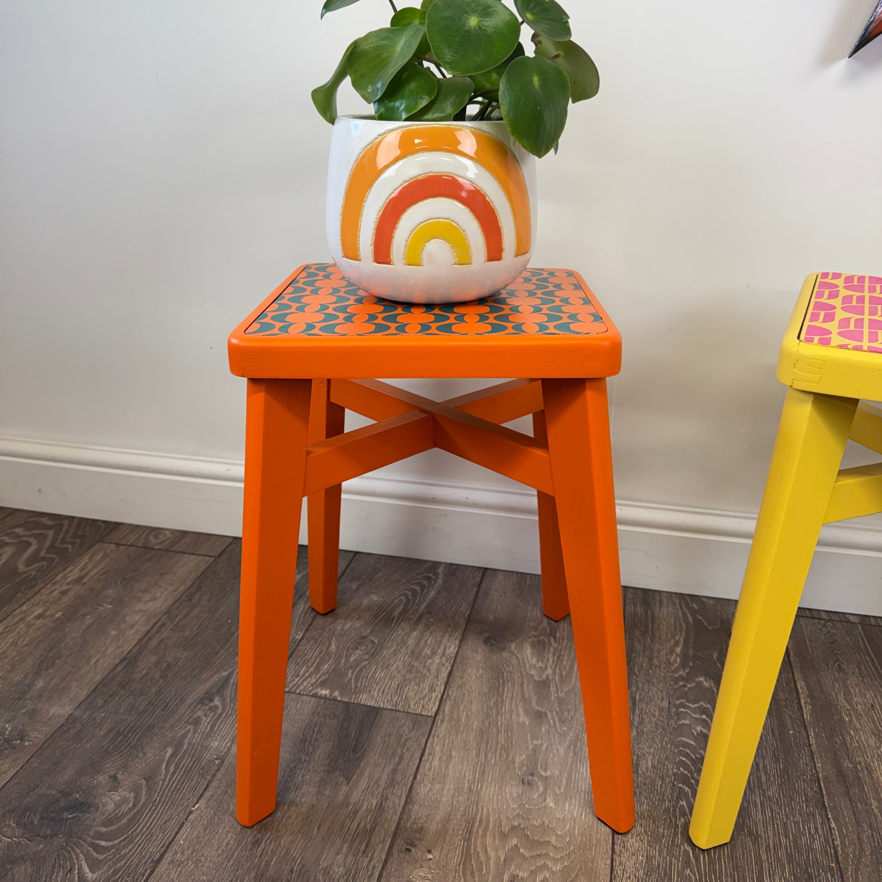 An orange wooden stool with a patterned top holding a potted green plant in a white pot with an orange and yellow sunburst design, set against a white wall and dark wood floor.
