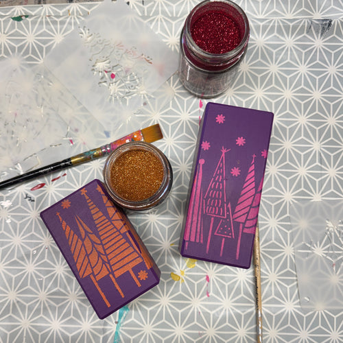 Two purple rectangular candle holders with pink christmas tree illustrations are arranged on a patterned surface alongside glitter jars, paintbrushes, and translucent craft sheets.