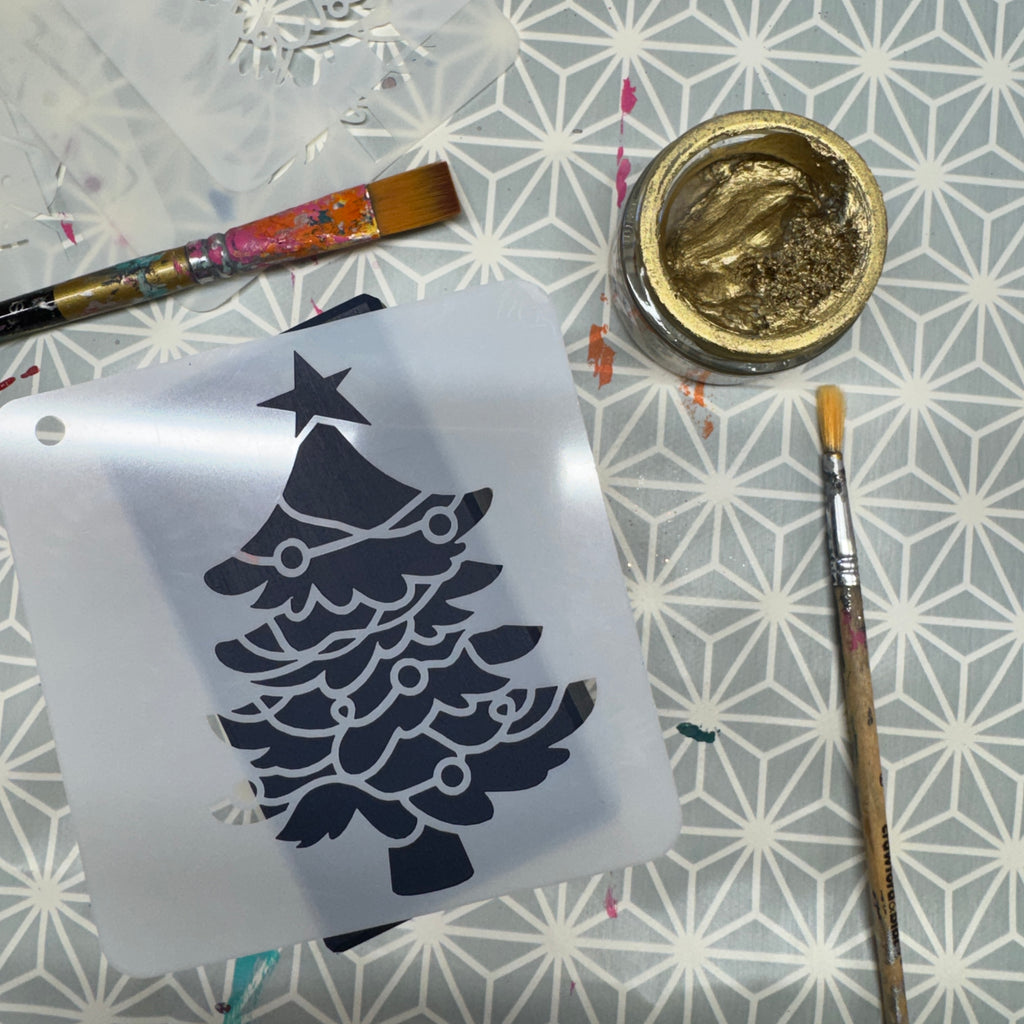 A stencil of a Christmas tree with ornaments lies on a patterned gray and white surface, surrounded by paintbrushes, a small jar of gold glitter paint, and a piece of white fabric.
