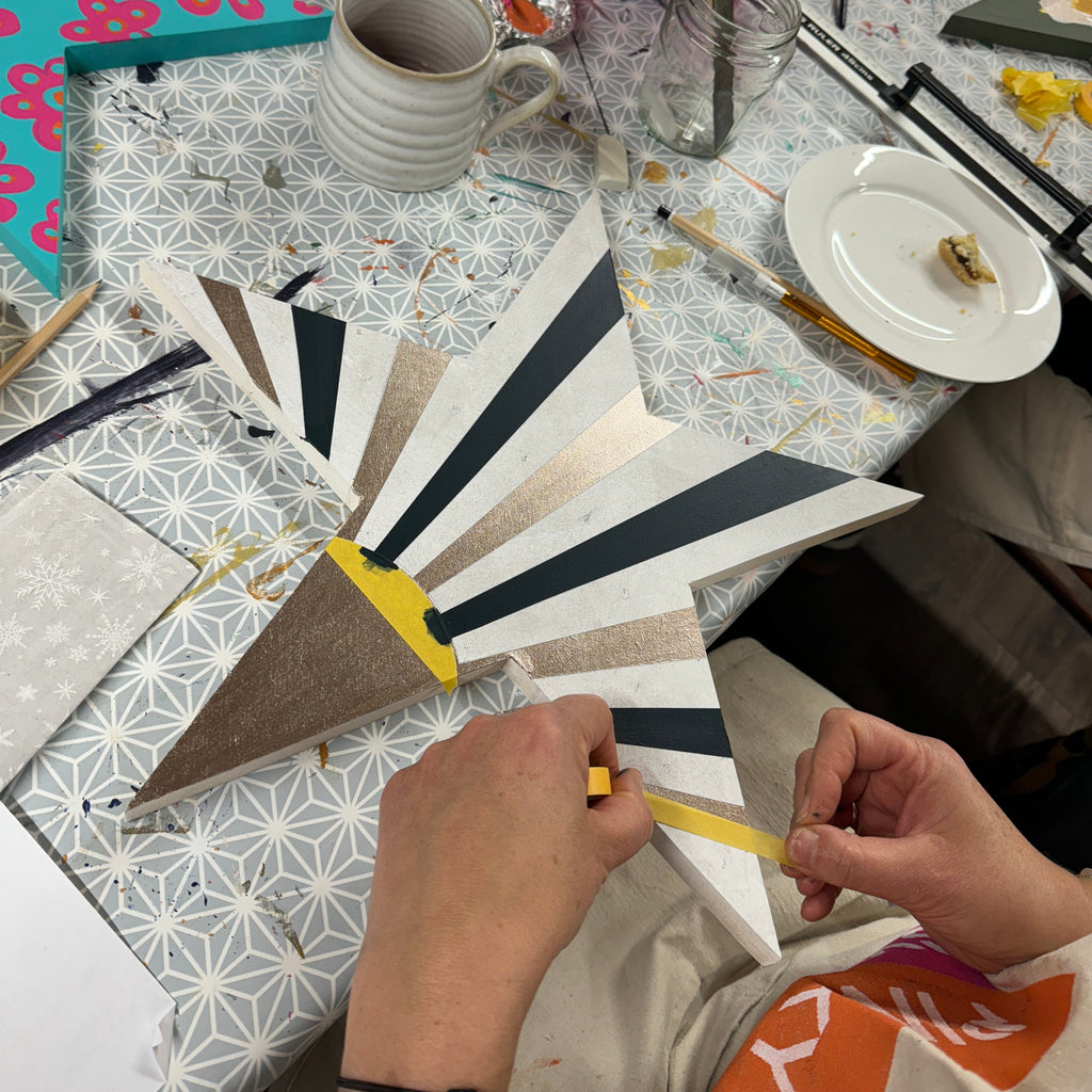 A person's hands are shown a large wooden christmas star on a busy, decorated craft table. The star features alternating black and white striped segments, a brown segment, and yellow accents.