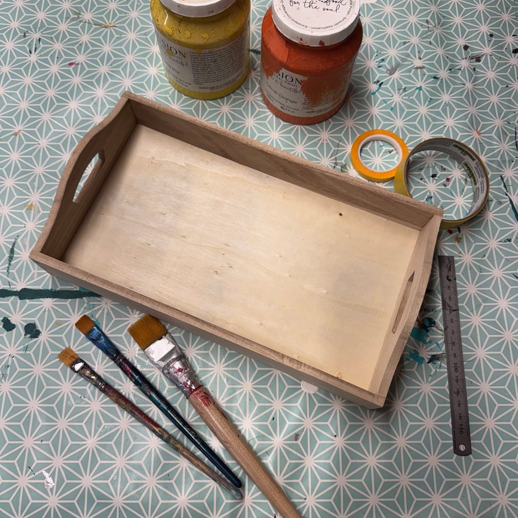 Wooden tray with paintbrushes, jars of paint, and tape on a patterned surface