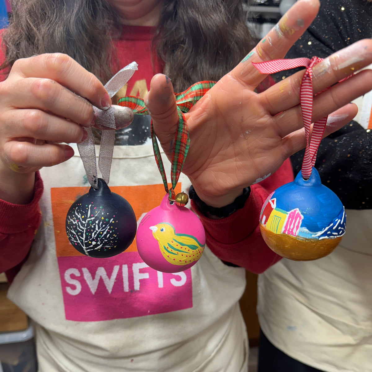 A person holding three hand-painted glass ornaments in both hands, wearing a white apron with 'SWIFTS' printed on it. The ornaments are decorated with a black tree design, a pink bird with a yellow beak, and a blue beach scene with a pink boat.