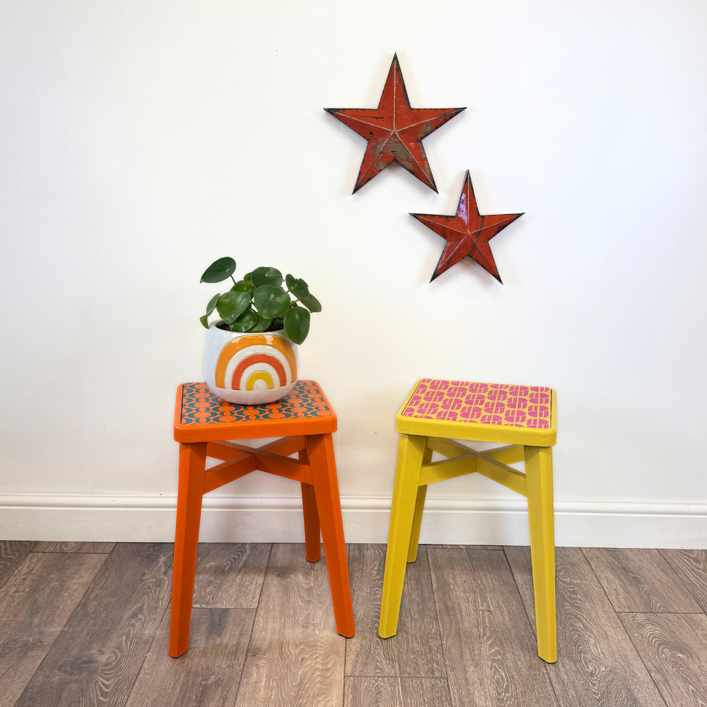 Two colorful wooden stools in orange and yellow, decorated with patterned seats, standing on a wood floor against a white wall with two red star decorations.